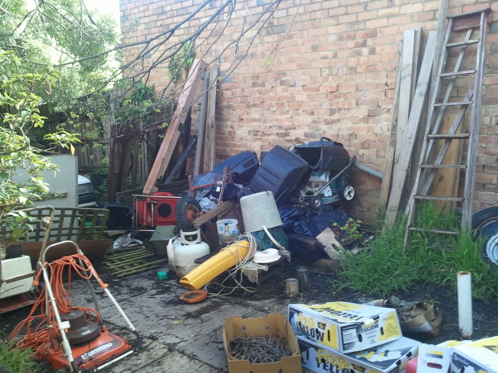 An outdoor area cluttered with a large accumulation of miscellaneous items against a brick wall, including old electronics, propane tanks, a ladder, cardboard boxes, and scrap metal. The scene illustrates conditions that may be addressed through public health nuisance or housing code enforcement.